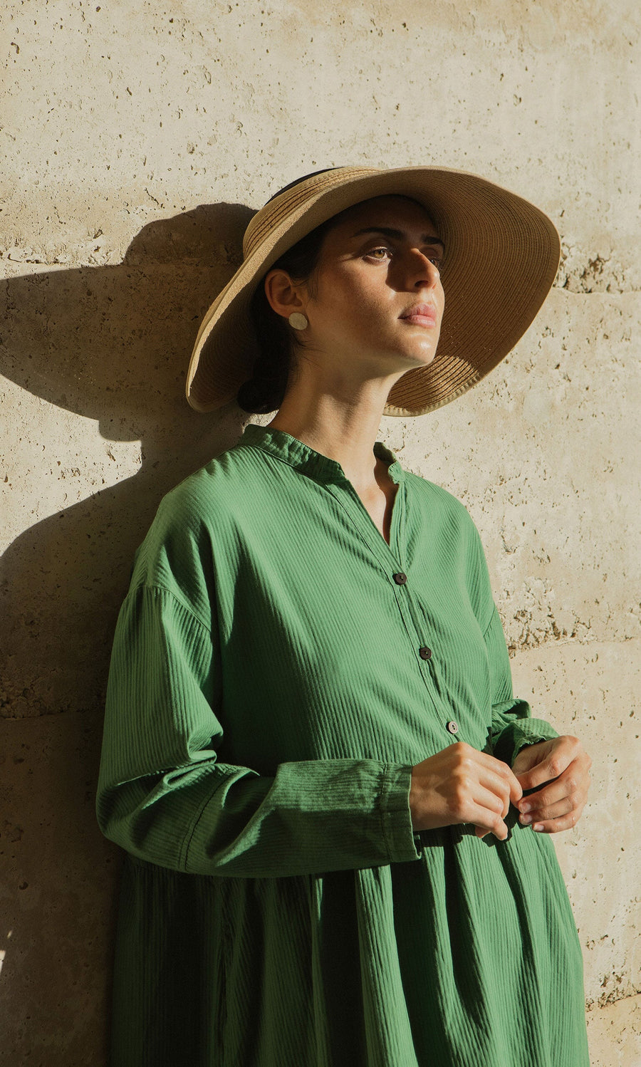 Plümo- A woman in the Marisol Dress with bracelet-length sleeves stands by a sunlit stone wall, gazing thoughtfully upward.