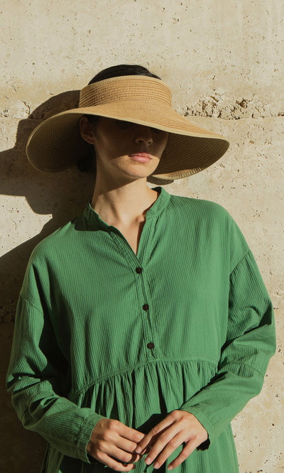 Plümo- Woman in a green dress wears the Miami Sun Visor, standing by a sunlit beige wall with her face partially in shadow.