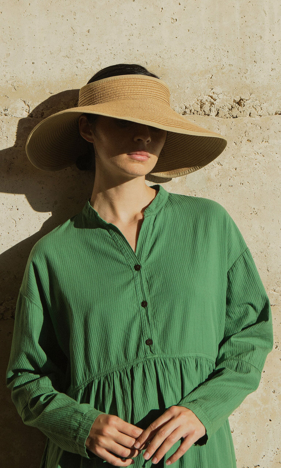 Plümo- Woman in a green dress wears the Miami Sun Visor, standing by a sunlit beige wall with her face partially in shadow.