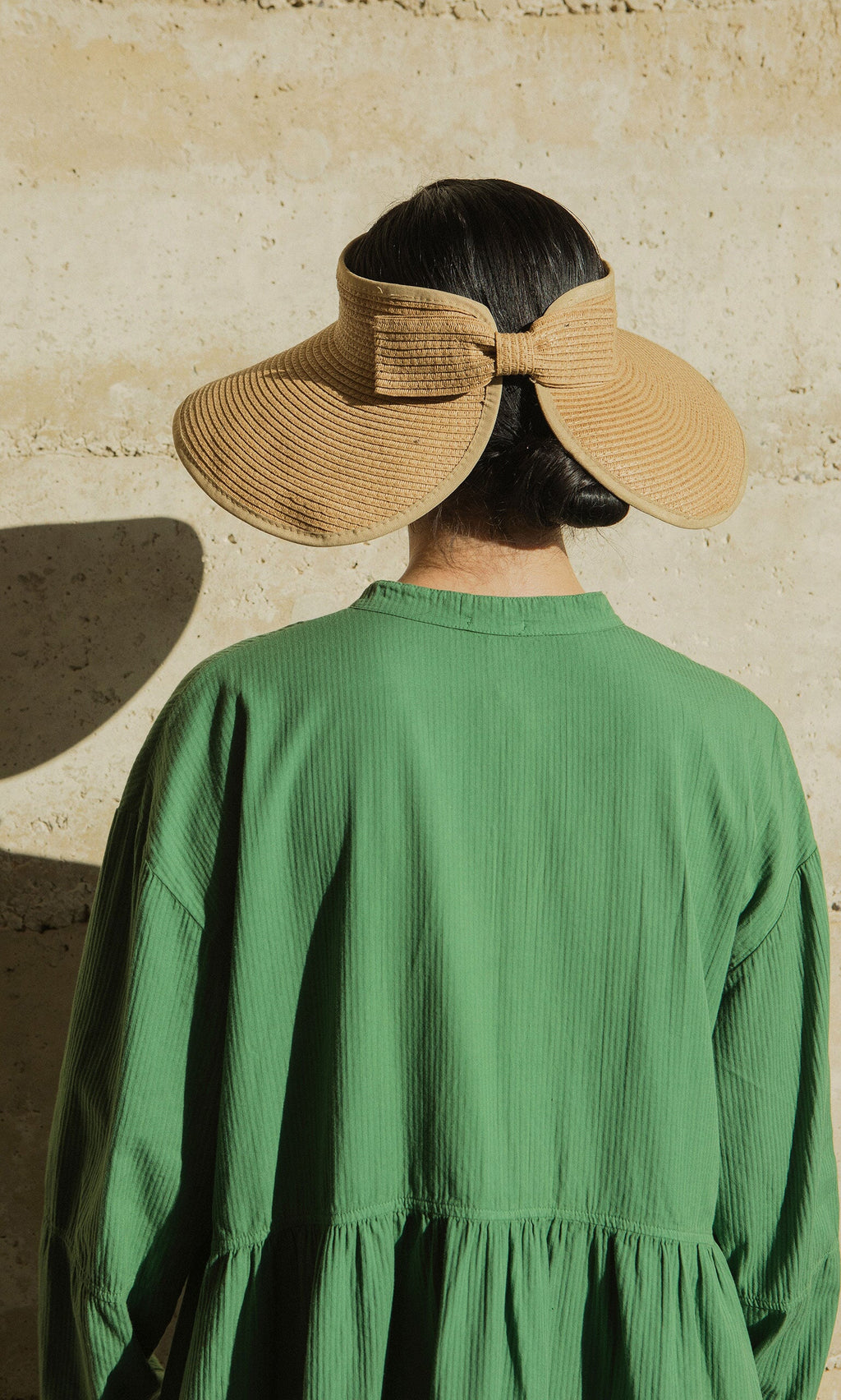 Plümo- Person in a green dress facing away, wearing the Miami Sun Visor with a back bow, against a beige wall.