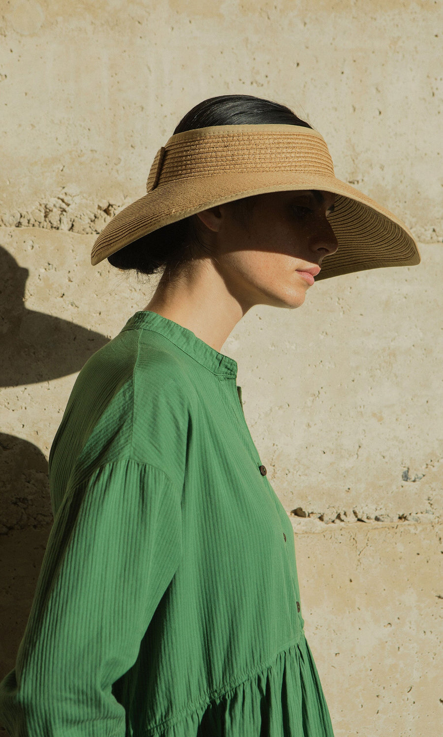 Plümo- Woman in a green dress wears the Miami Sun Visor, standing in sunlight by a textured beige wall.