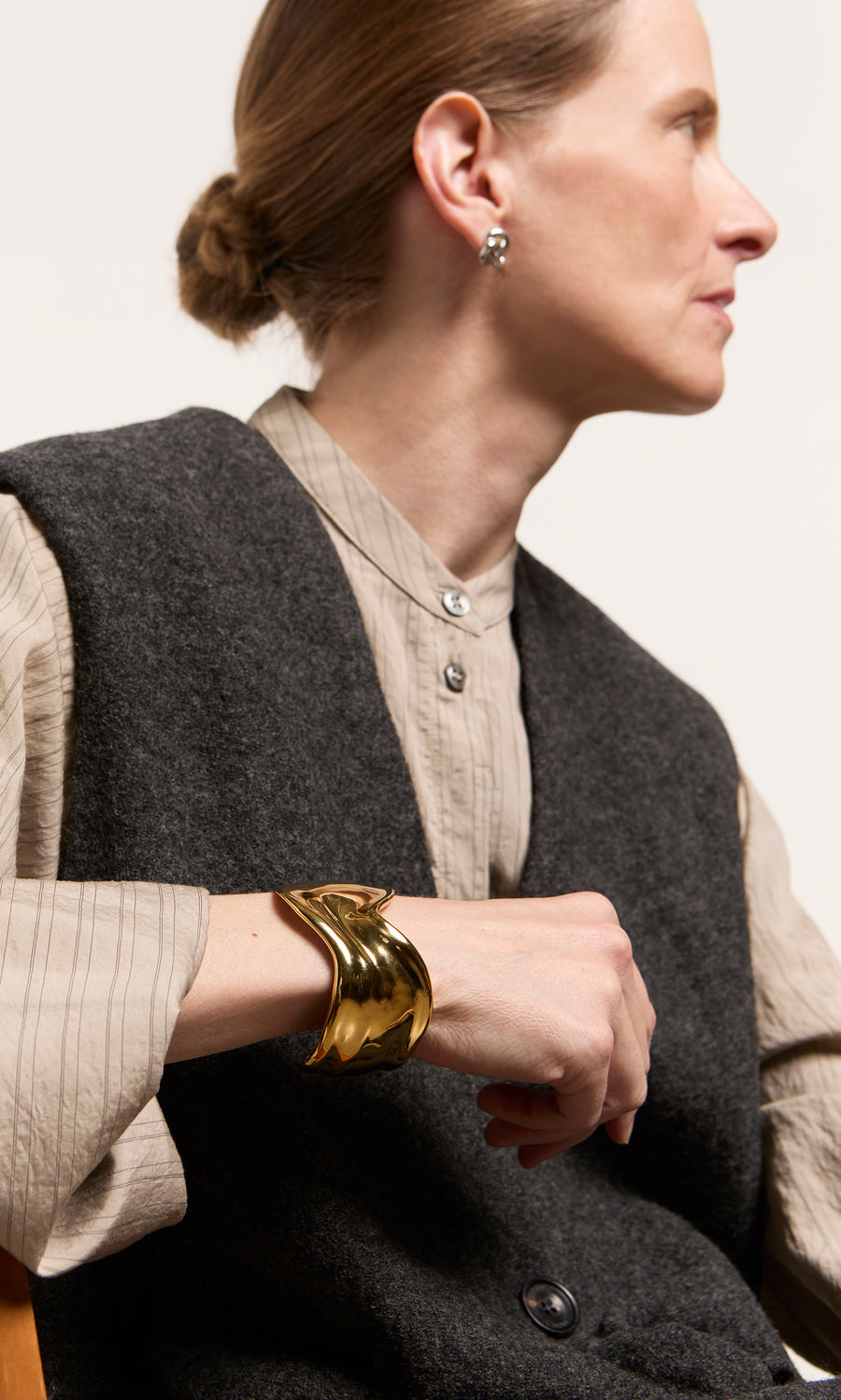 Plümo  Woman in profile wearing the Mira Cuff, gray vest, beige shirt, and earrings, seated on a wooden chair.