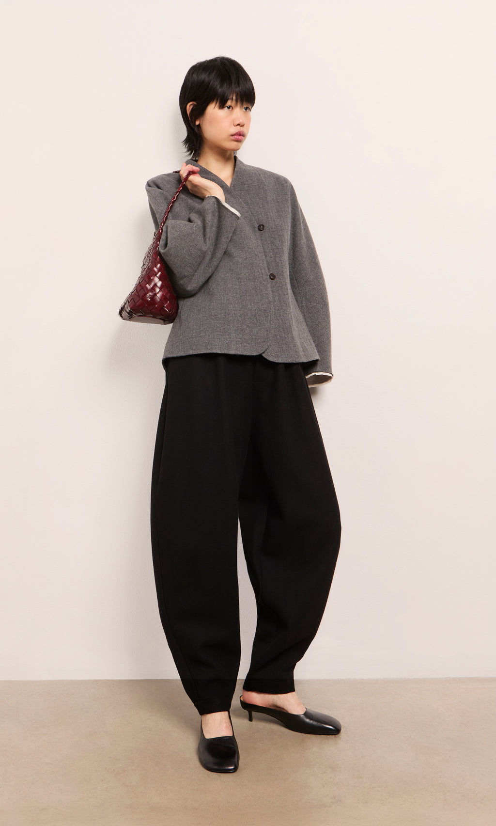 Plümo  Woman in a gray blazer, Circle Pants - Boiled Black, black heels, and a maroon bag stands by a plain wall.