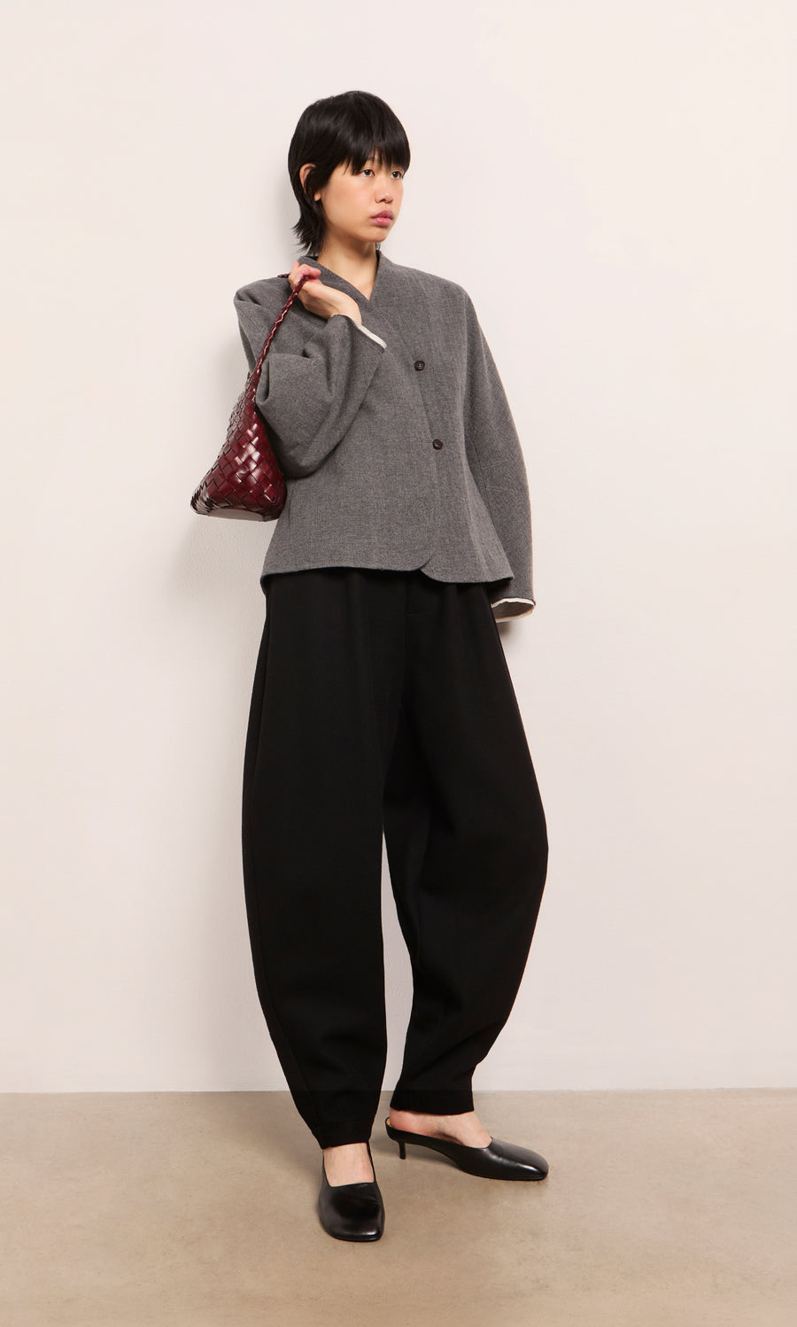 Plümo  Woman in a gray blazer, Circle Pants - Boiled Black, black heels, and a maroon bag stands by a plain wall.