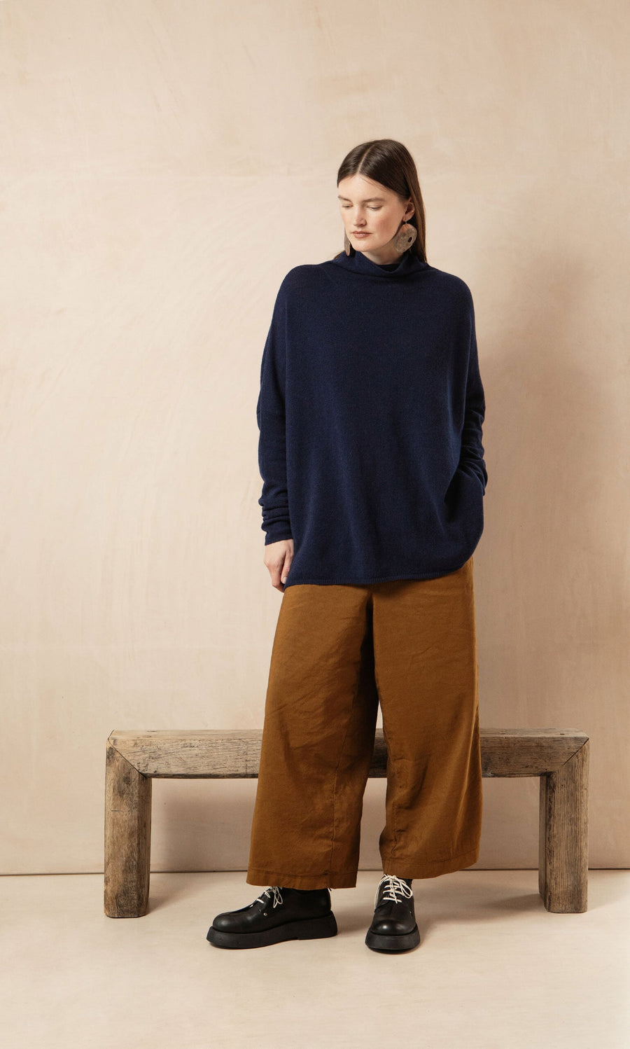 Plümo- A woman in a Navy Rollneck and wide brown pants stands by a rustic bench, looking down.