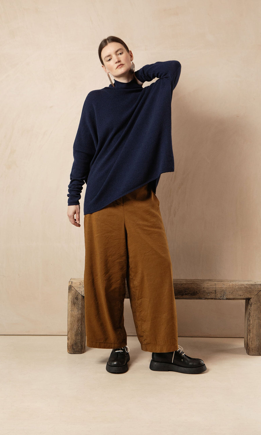 Plümo- Woman in a Navy Rollneck stands by a rustic bench against a beige background, styled with brown wide-leg pants.