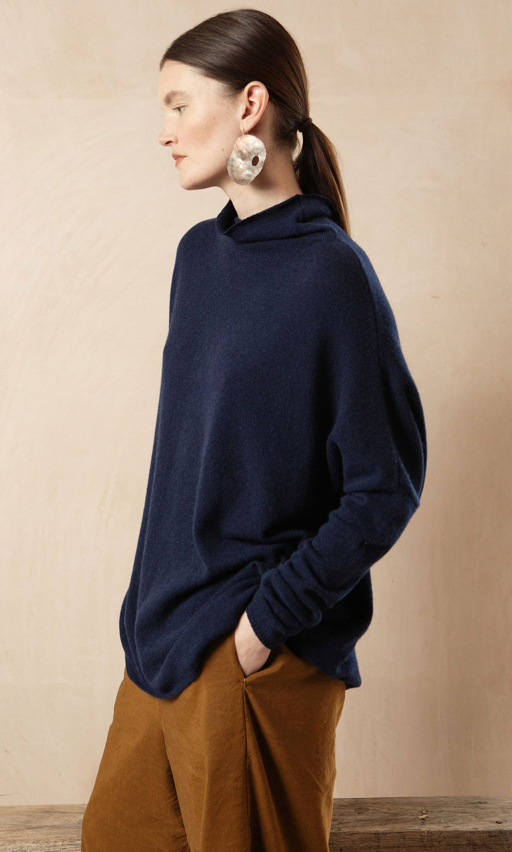 Plümo- Woman in a Navy Rollneck sweater and brown pants stands sideways with hand in pocket, wearing large earrings.
