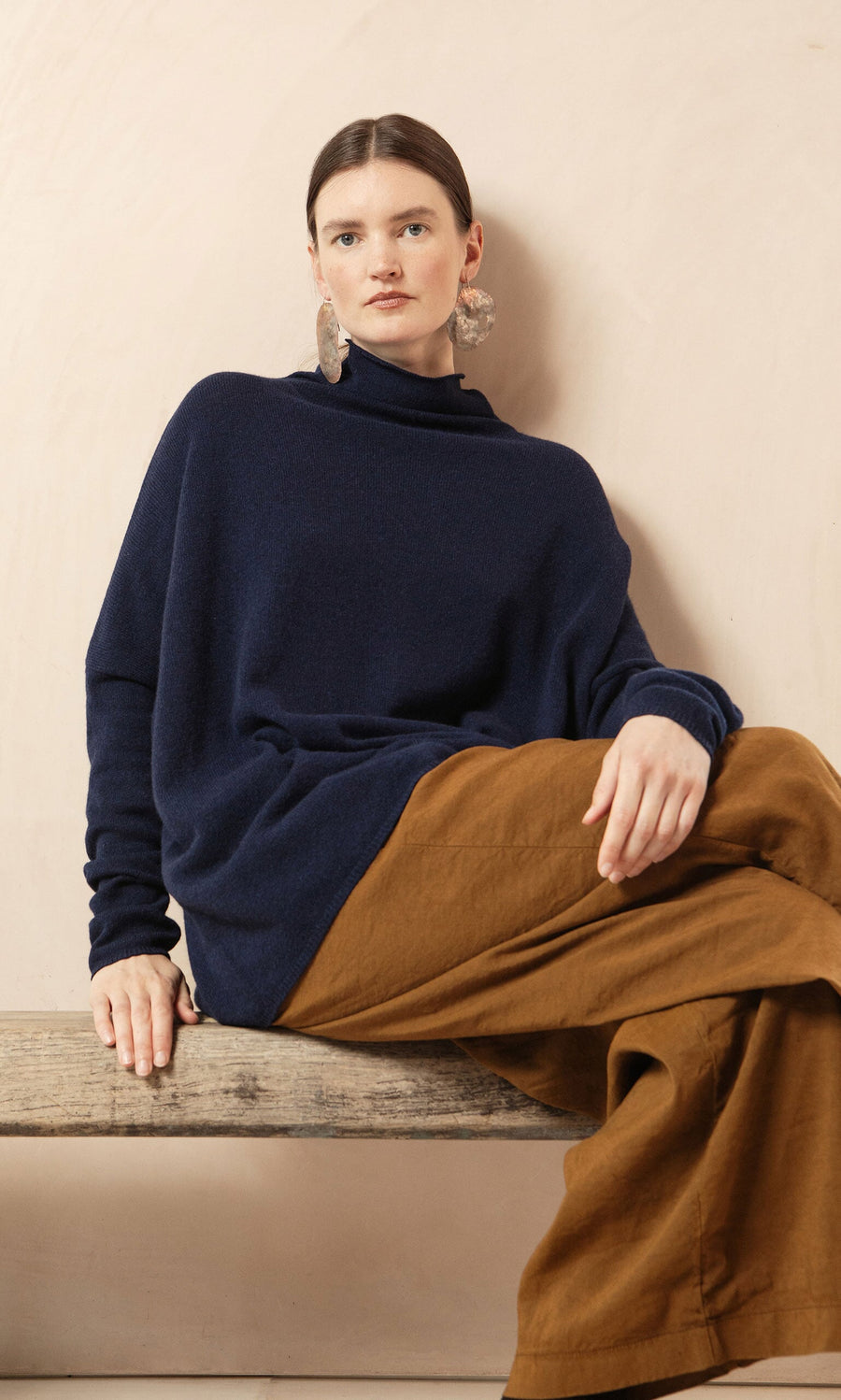 Plümo- Woman sitting on a bench in a relaxed fit Navy Rollneck, brown wide-leg pants, and large statement earrings.