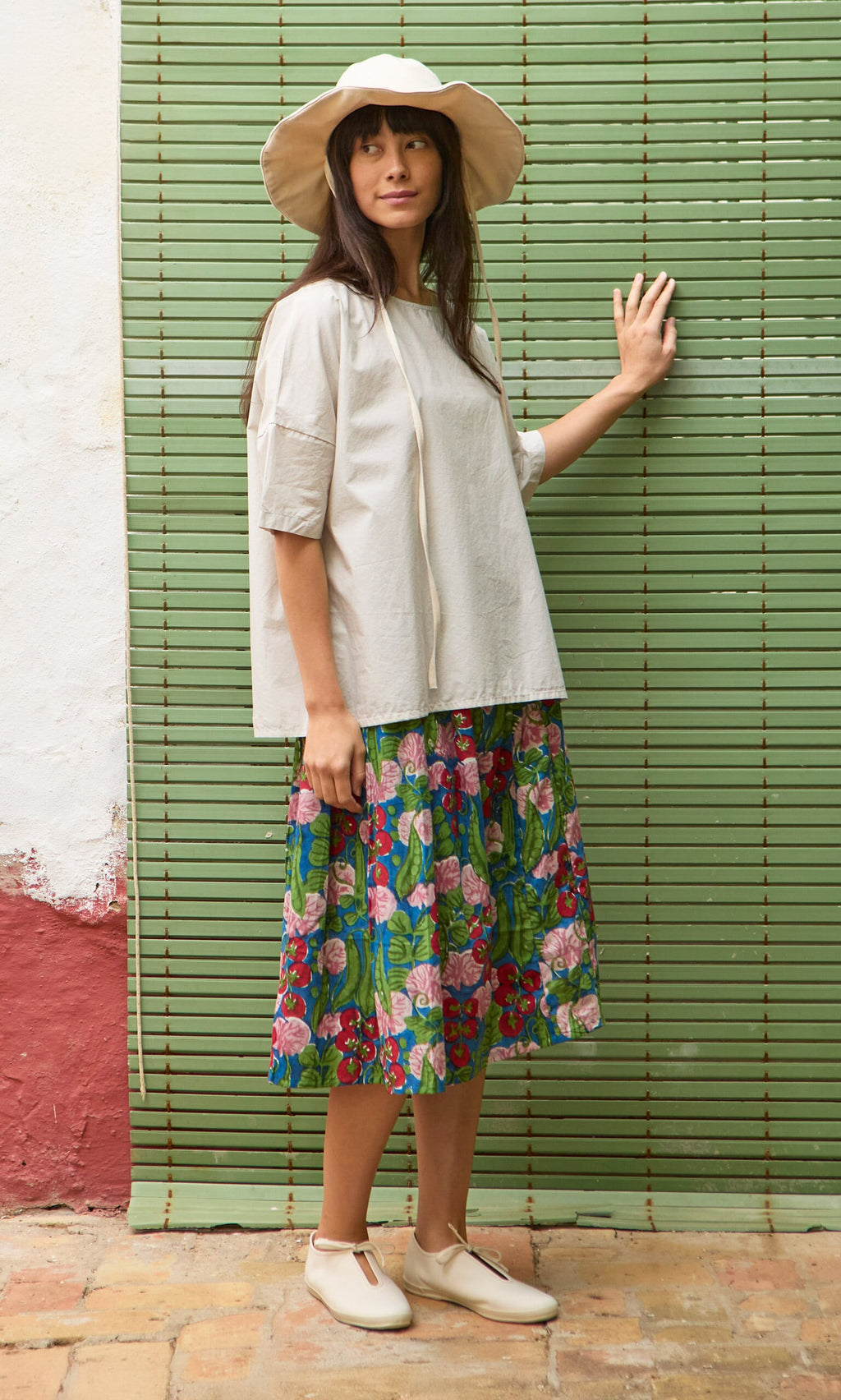 Plümo- Woman in a wide-brim hat, Nellie Tee, floral skirt, and beige shoes stands by a green bamboo blind.