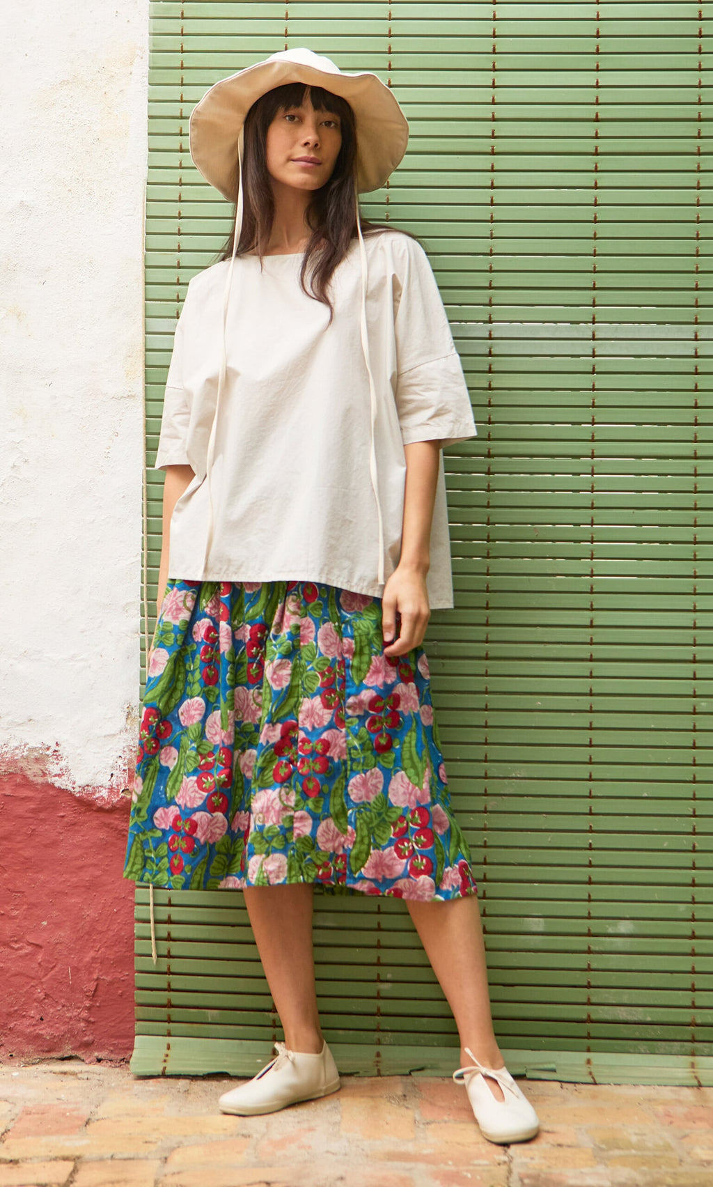 Plümo- Woman in a floppy hat, Nellie Tee, and floral skirt stands by a green panel wall.