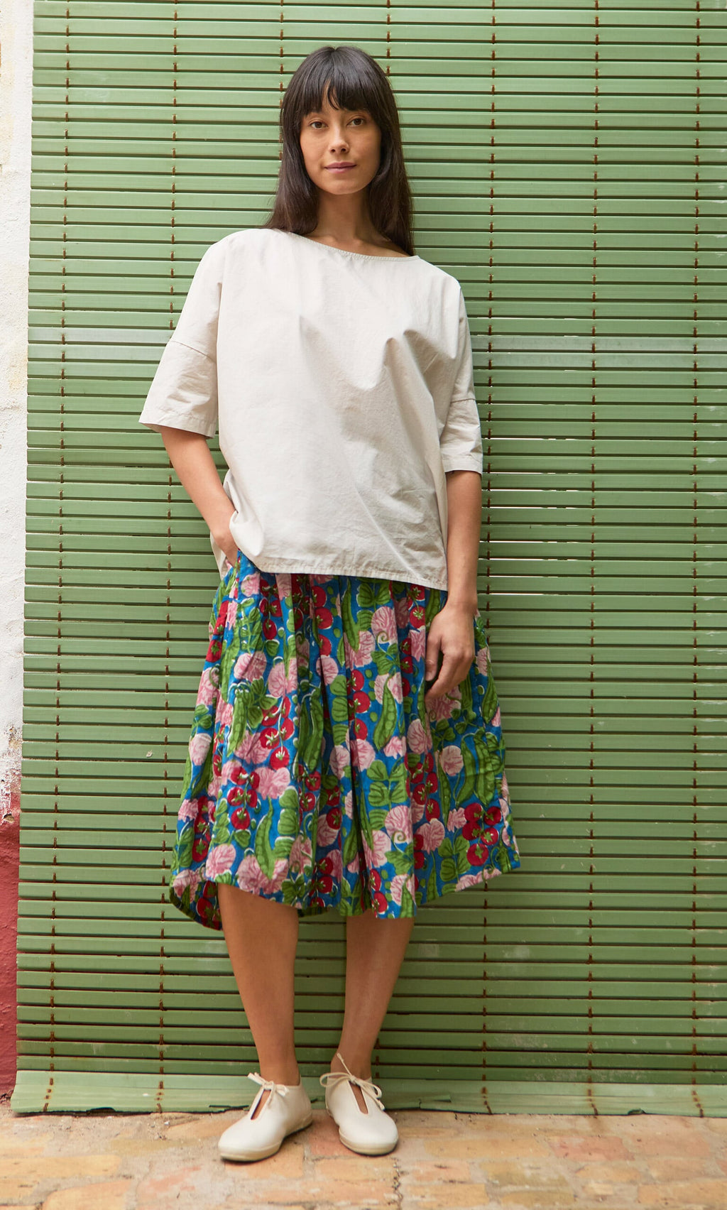 Plümo- Woman in a Nellie Tee, colorful floral skirt, and white shoes stands before a green wall.
