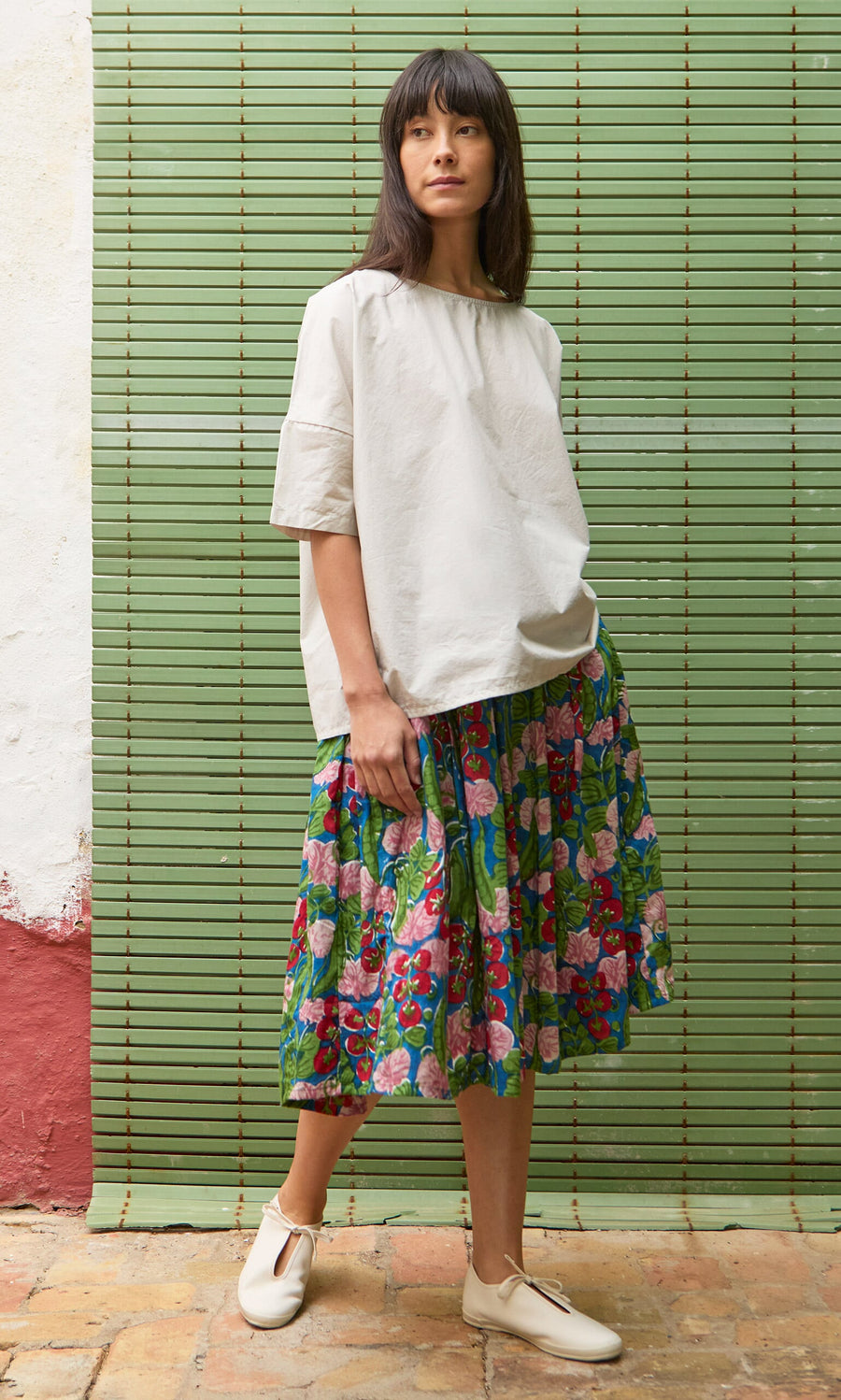 Plümo- Woman in a boxy Nellie Tee and floral skirt poses on a tiled floor before a green slatted wall.