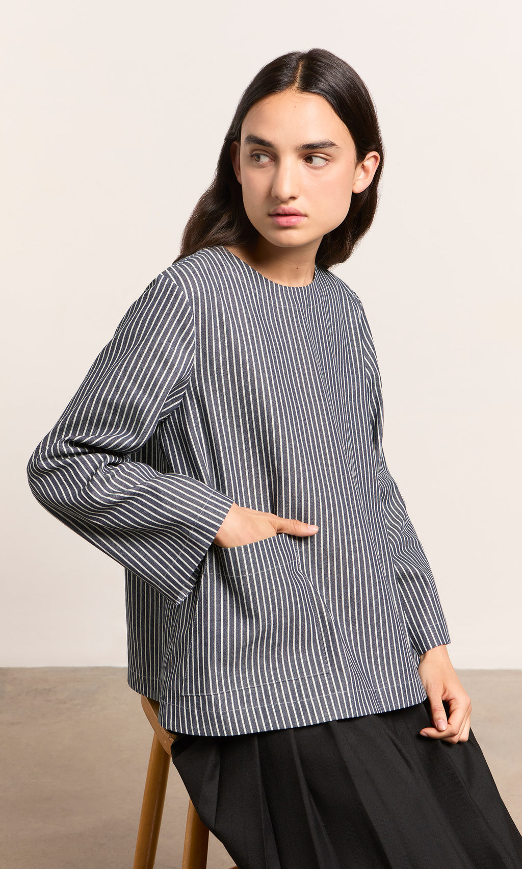 Plümo  Person wears Norfolk Top, sitting on a stool, looking to the side with hand in pocket.