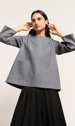 Plümo  Person in a Norfolk Top and black skirt poses with hands near head against a plain background.