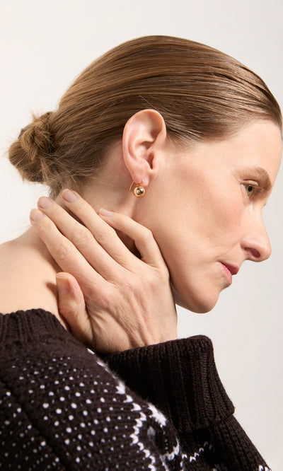 Plümo  Woman with brown hair in a bun, black sweater, wearing Ora Earrings, touches her neck and jawline.