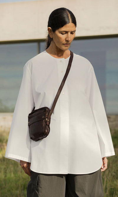 Plümo- Person with long dark hair in a loose white shirt carries the Orton Parcel Bag, standing outdoors by a building.
