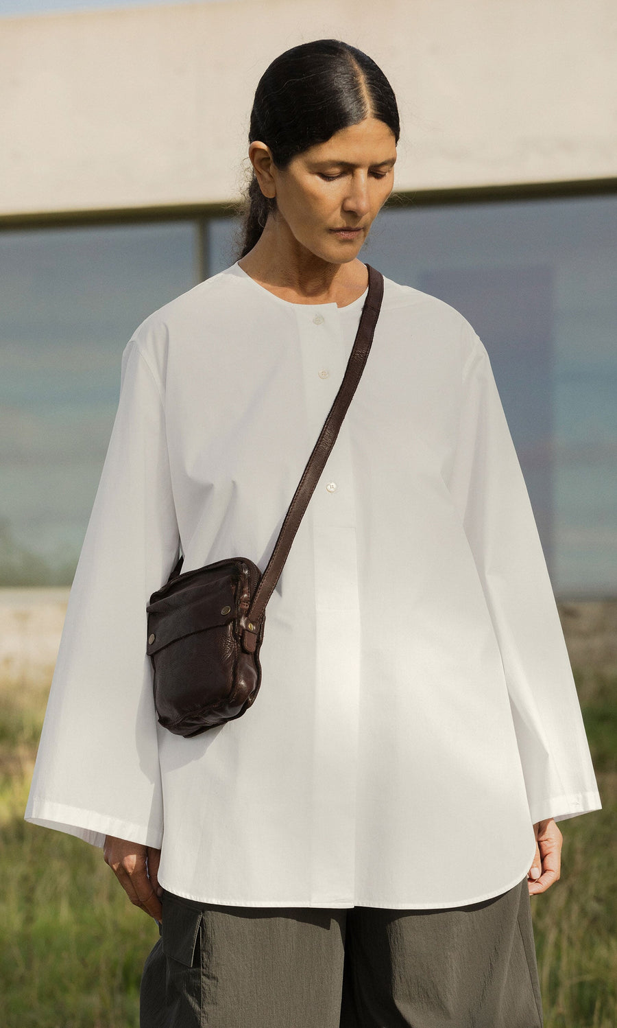 Plümo- Person with long dark hair in a loose white shirt carries the Orton Parcel Bag, standing outdoors by a building.