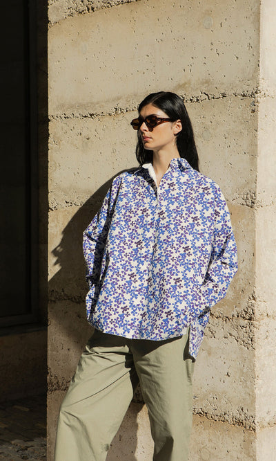 Plümo- Woman in Hannoh Wessel sunglasses and an Osterley Shirt stands by a sunlit textured stone wall.