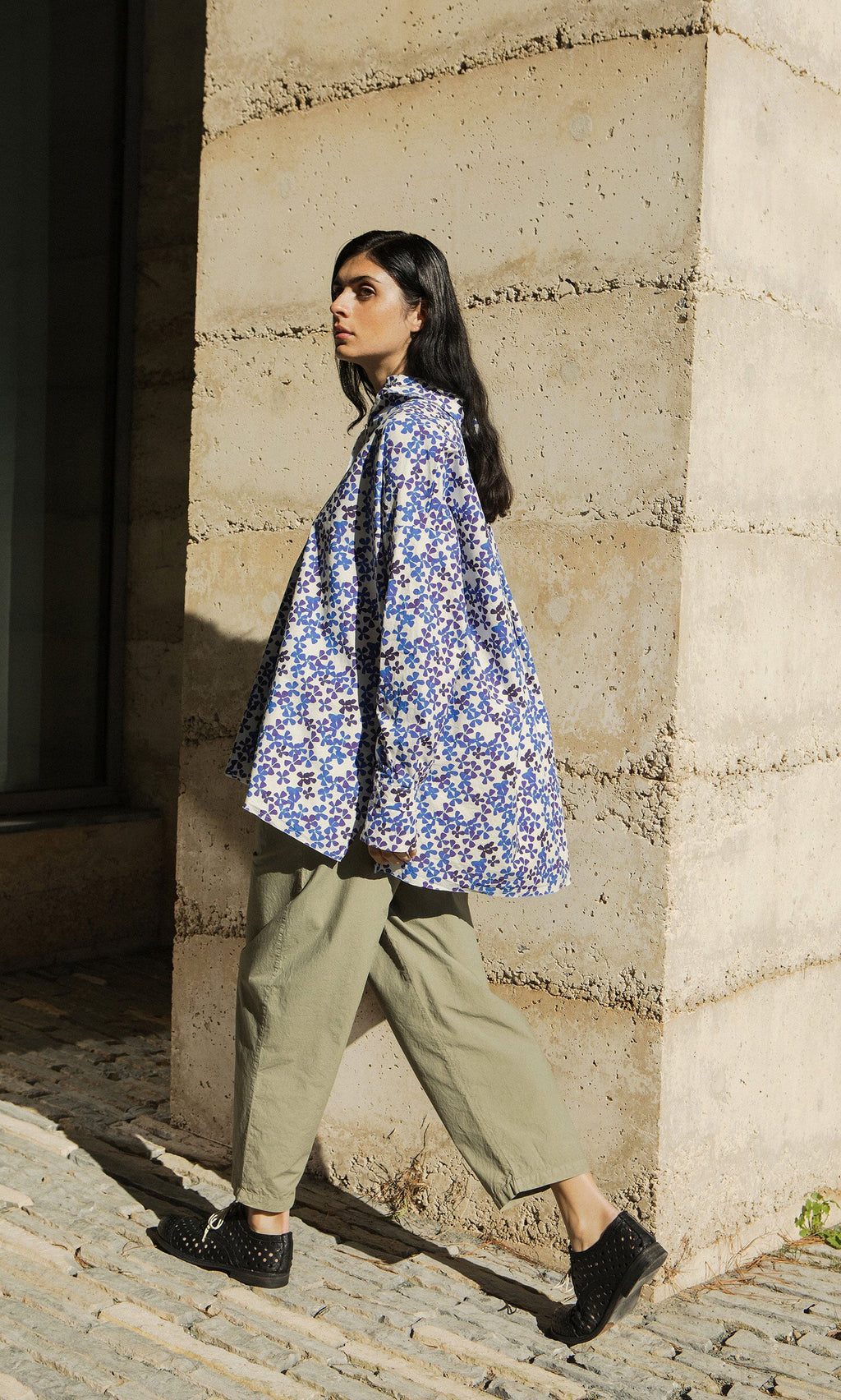 Plümo- Woman in an Osterley Shirt and khaki pants walks on a cobblestone path by a concrete wall.
