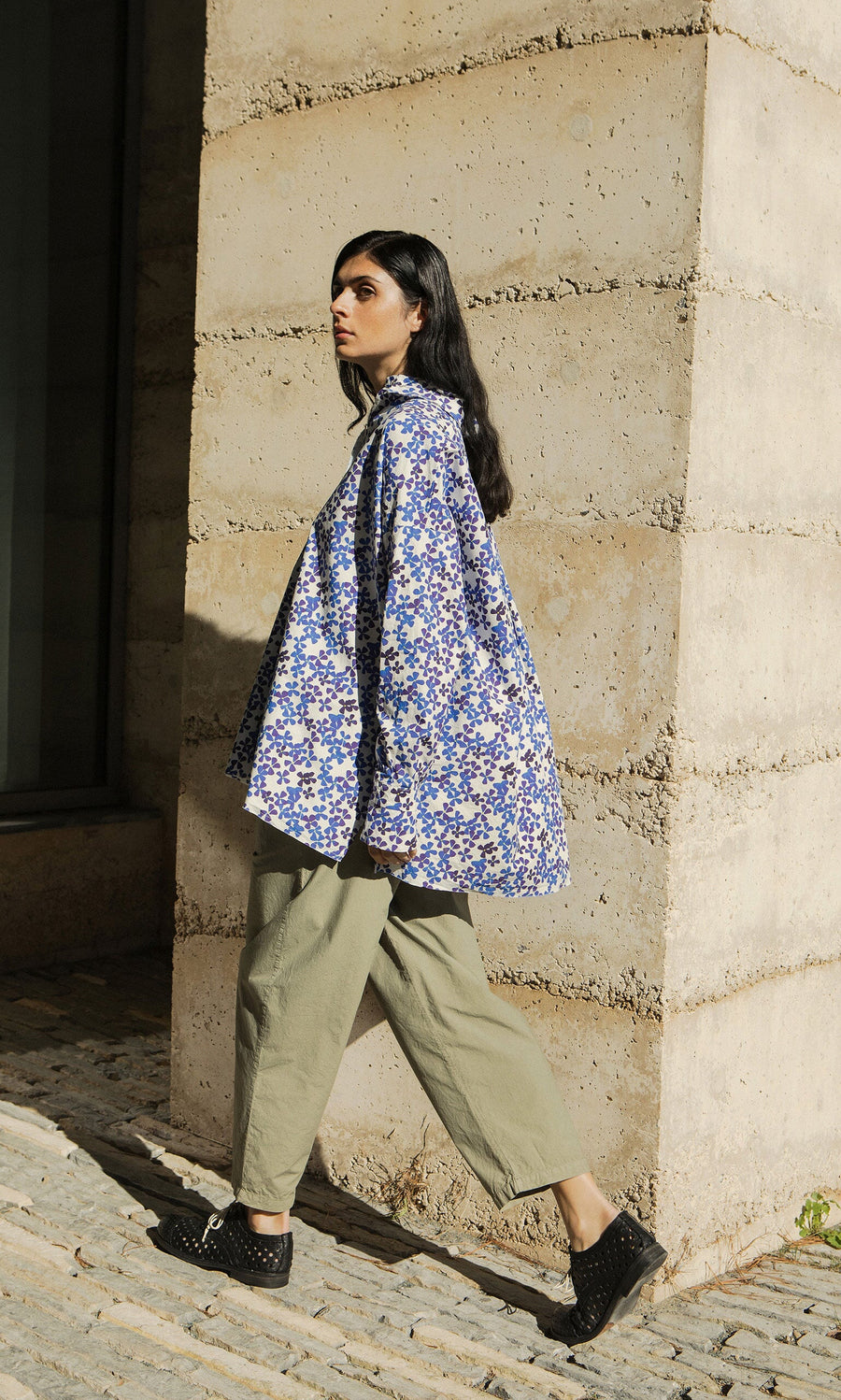 Plümo- Woman in an Osterley Shirt and khaki pants walks on a cobblestone path by a concrete wall.
