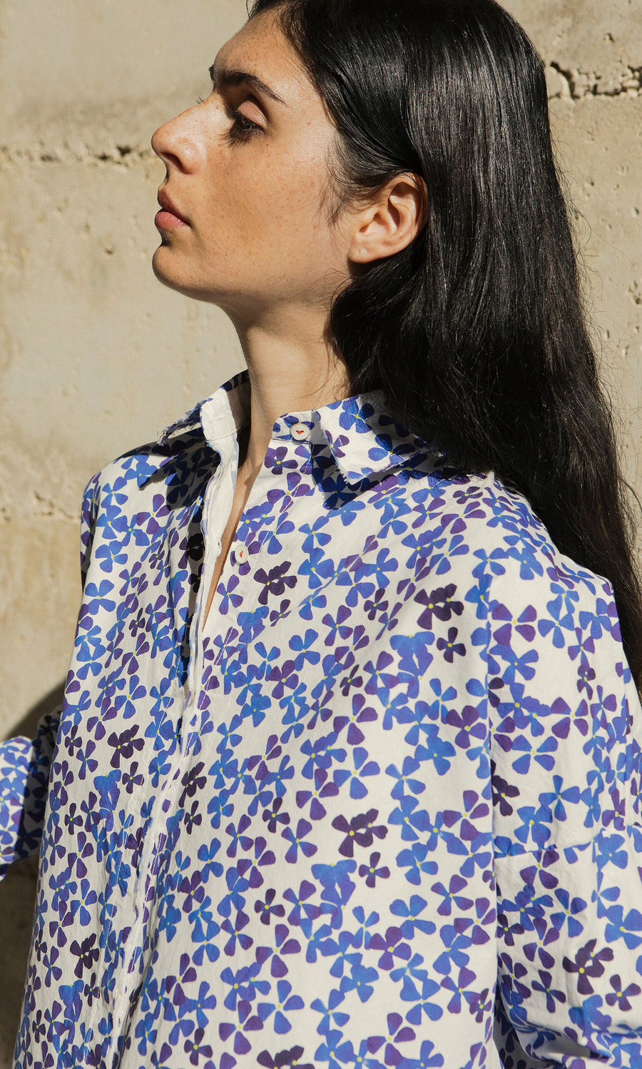 Plümo- Woman with long dark hair in a violet blue Osterley Shirt by Hannoh Wessel, standing against a beige wall.