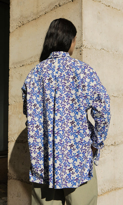 Plümo- Person with long dark hair wears an Osterley Shirt, standing by a stone wall.