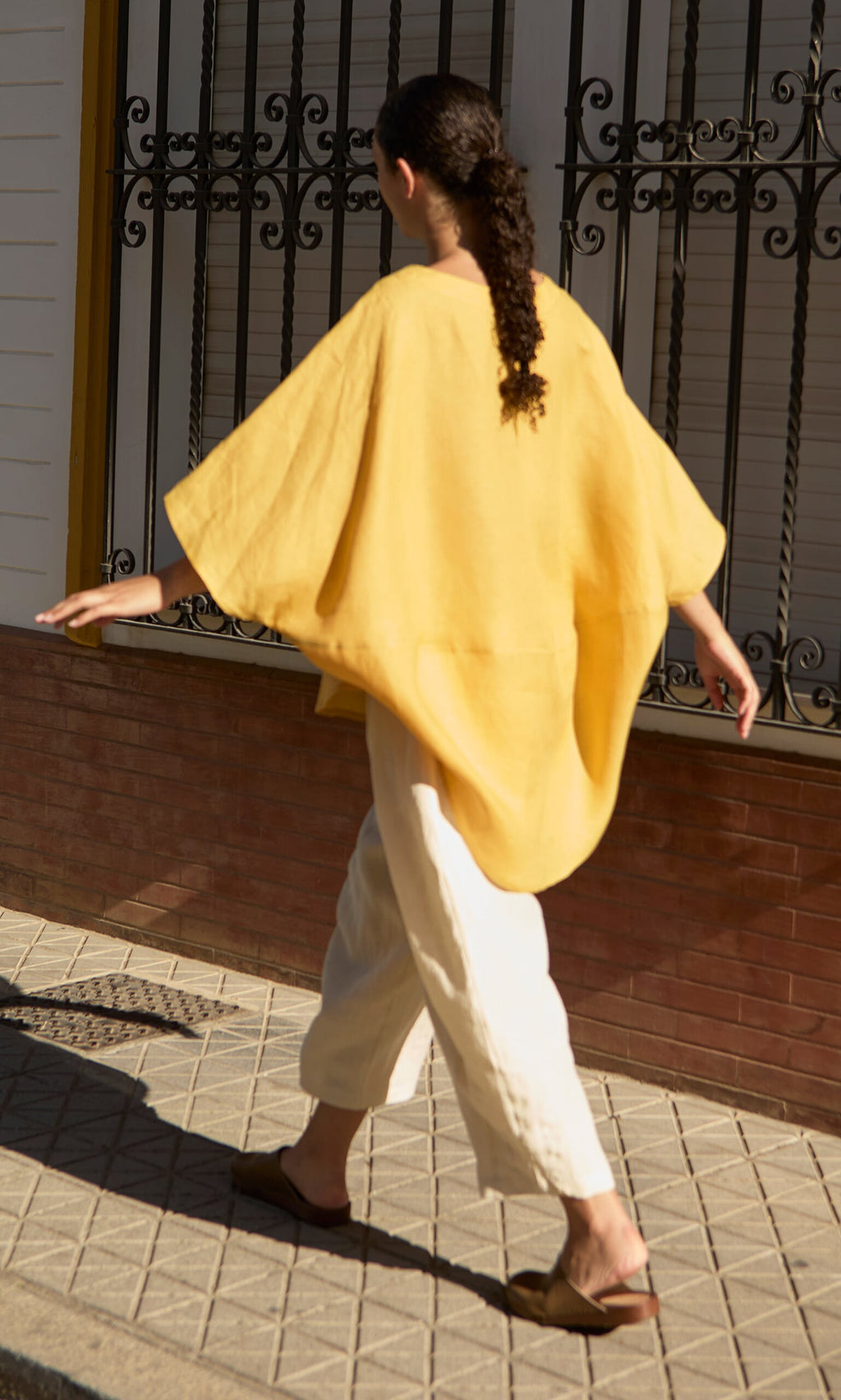 Plümo- Someone in a yellow poncho and the Otilia Blouse - Sunny walks on a sunlit sidewalk by barred windows.