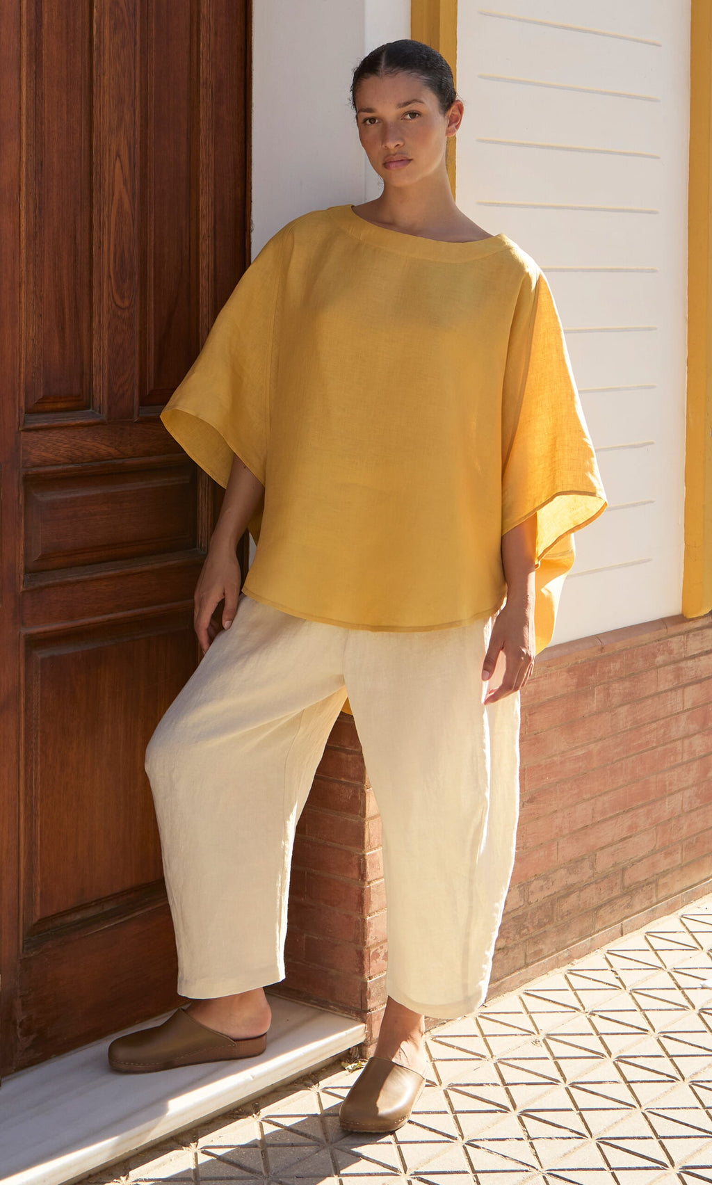 Plümo- Woman wearing the Otilia Blouse - Sunny and cream pants stands by a wooden door on a sunny day, facing the camera.