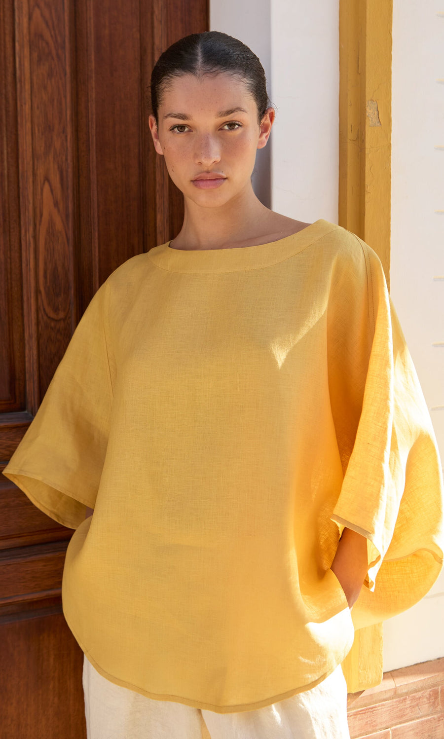 Plümo- A woman wears the Otilia Blouse - Sunny in front of a wooden door and yellow-trimmed wall, facing the camera.