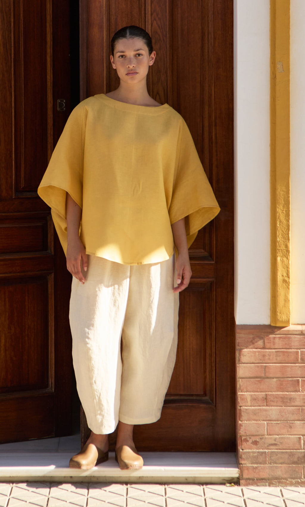 Plümo- Person wears the Otilia Blouse - Sunny and cream pants in front of a wooden door and yellow-trimmed wall.