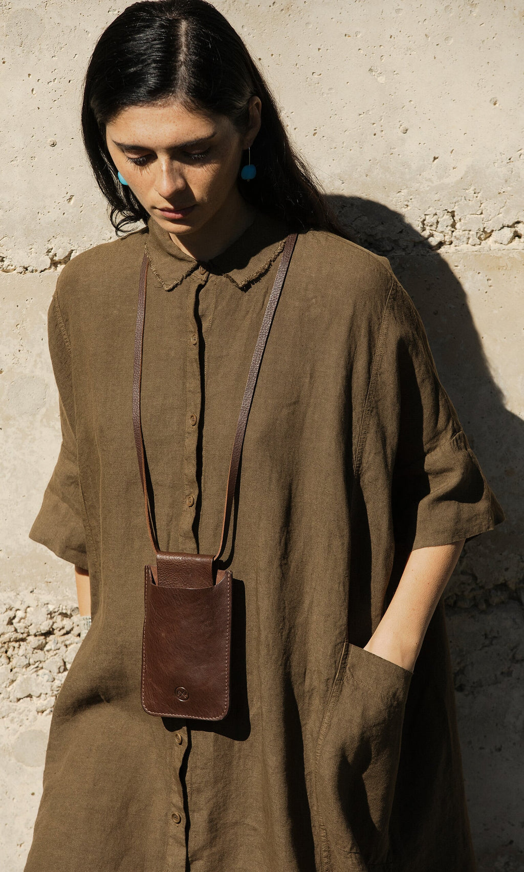 Plümo- Woman in a loose brown dress wears the Purdy Bag, a vegetable-dyed leather pouch necklace, by a concrete wall.