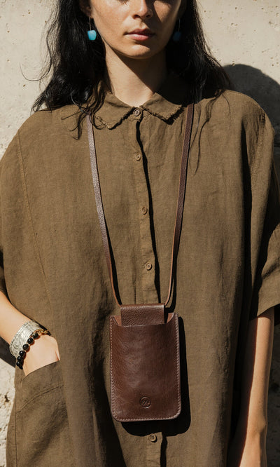 Plümo- Woman in a loose brown shirt with the Purdy Bag crossbody, standing against a light textured wall.