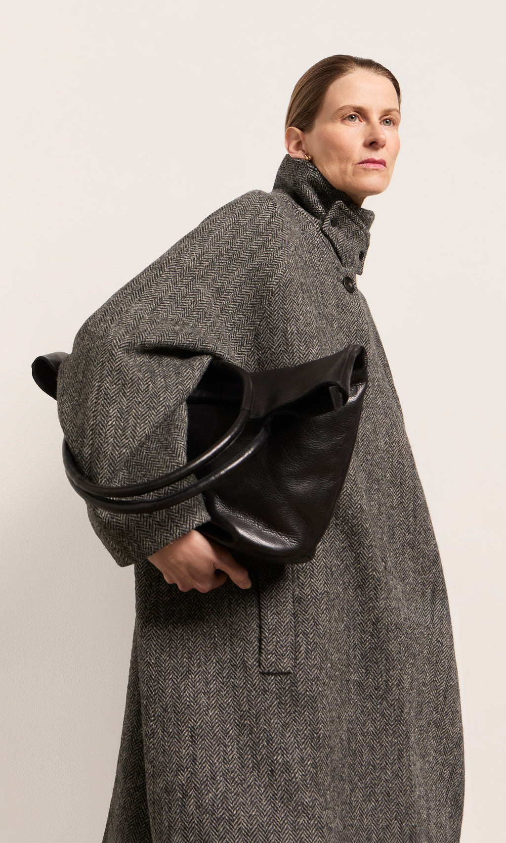 Plümo  Woman in a gray coat holds the Oval Shoulder Bag in black leather, looking confidently to the side.