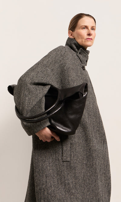 Plümo  Woman in a gray coat holds the Oval Shoulder Bag in black leather, looking confidently to the side.