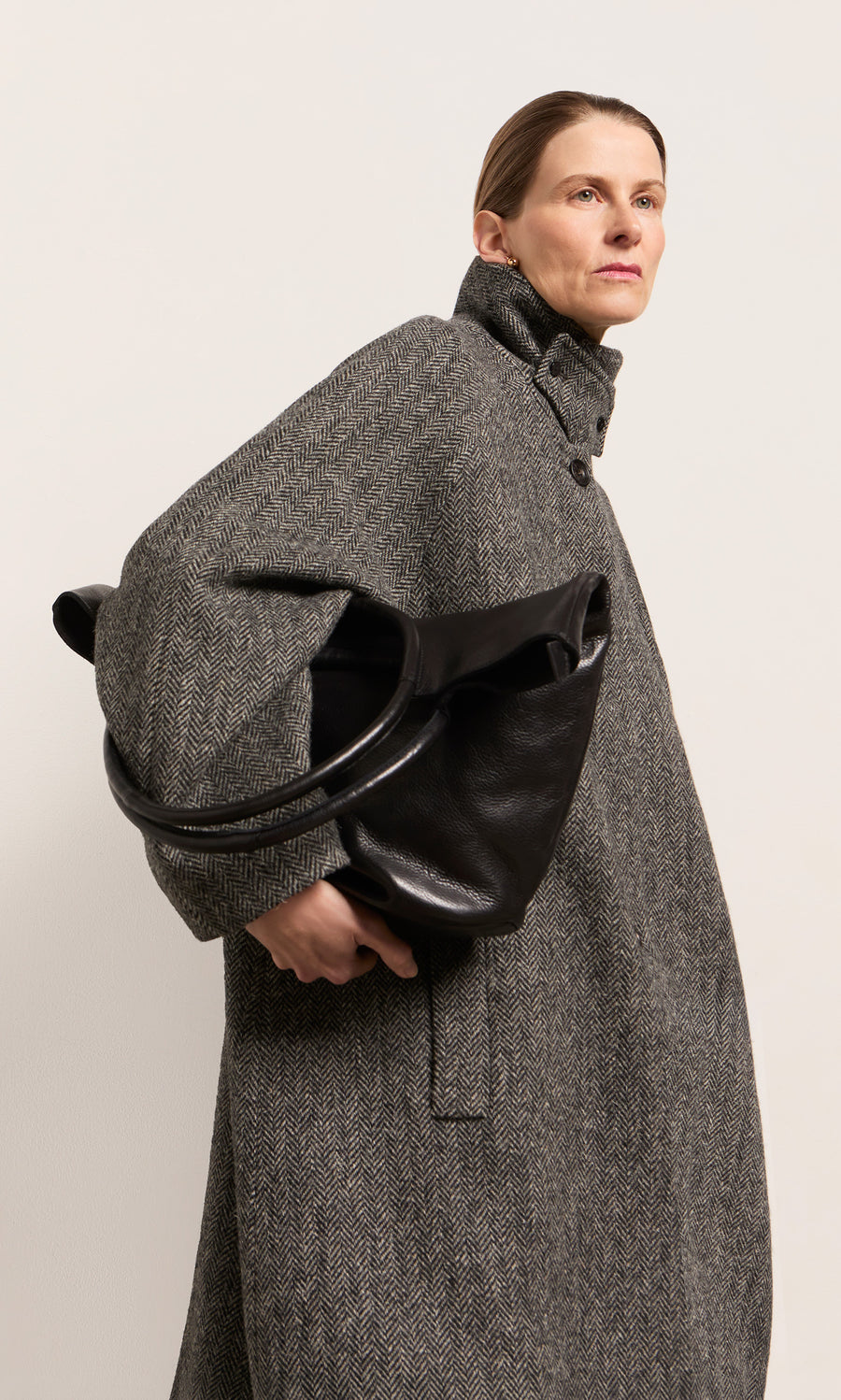 Plümo  Woman in a gray coat holds the Oval Shoulder Bag in black leather, looking confidently to the side.