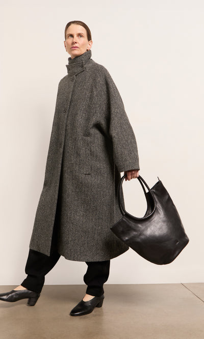 Plümo  Person in a long grey coat, black pants, holding an Oval Shoulder Bag, standing indoors.
