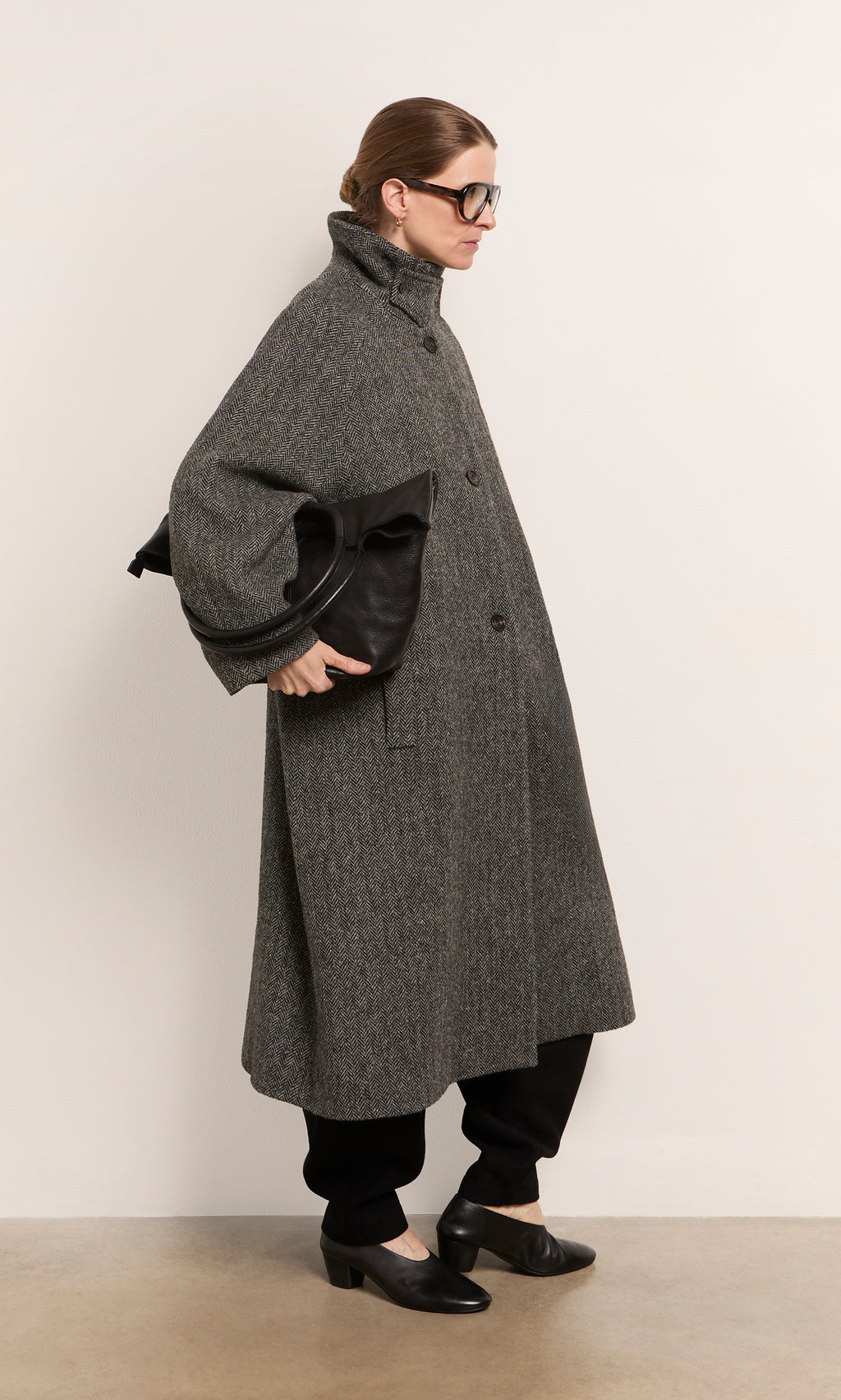 Plümo  Person in a long gray coat, black pants and shoes, holding an Oval Shoulder Bag, stands against a plain wall.