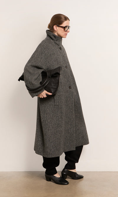 Plümo  Person in a long gray coat, black pants and shoes, holding an Oval Shoulder Bag, stands against a plain wall.