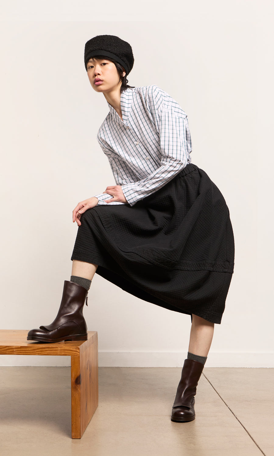 Plümo  Person in plaid shirt, black skirt, boots, and a handmade Perinne Beret poses with one leg up on a wooden bench.