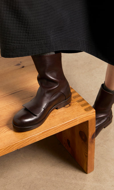 Plümo  A person in Over Boots and a black skirt stands with one foot on a wooden bench.