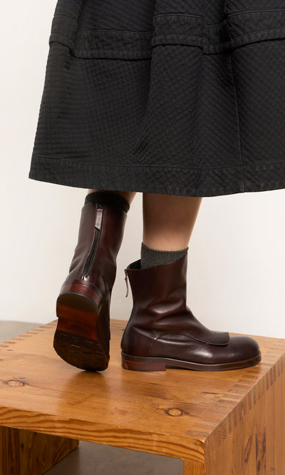 Plümo  Person in Over Boots—artisan-made in Italy, dark brown—gray socks, black skirt, stands on wooden table.