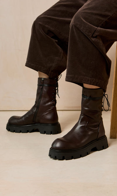 Plümo- Person wearing Paola Boots—artisan-crafted dark brown lace-ups with black chunky soles—sitting in brown pants on a wooden chair.