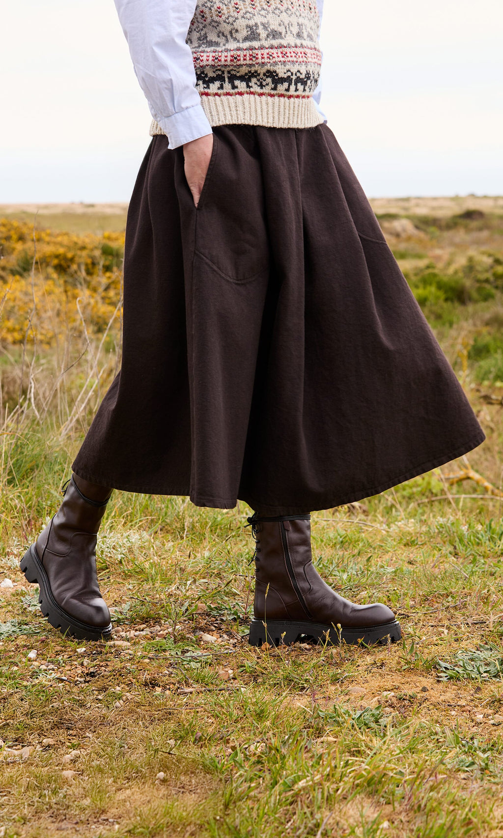 Plümo- Person outdoors on grassy terrain in the Allard Skirt, patterned sweater vest, white shirt, and brown boots.