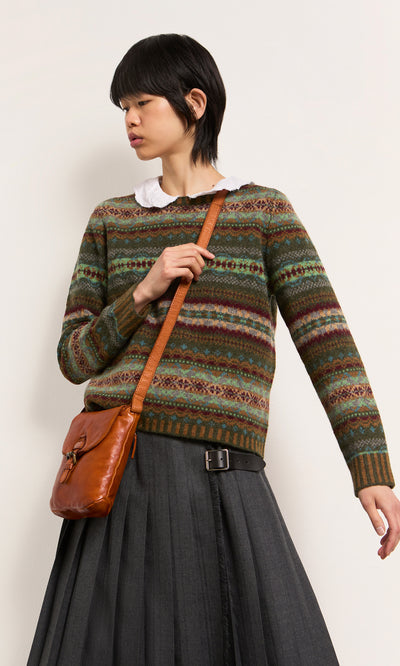 Plümo  Person in a patterned sweater, pleated gray skirt, and Parcel Bag - Chestnut stands against a plain background.
