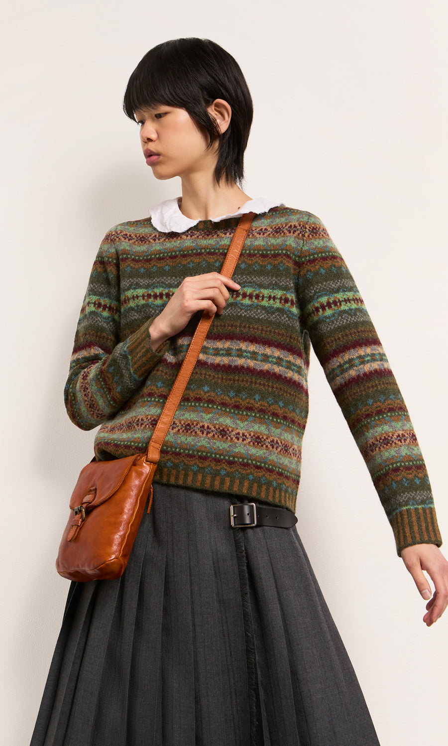 Plümo  Person in a patterned sweater, pleated gray skirt, and Parcel Bag - Chestnut stands against a plain background.
