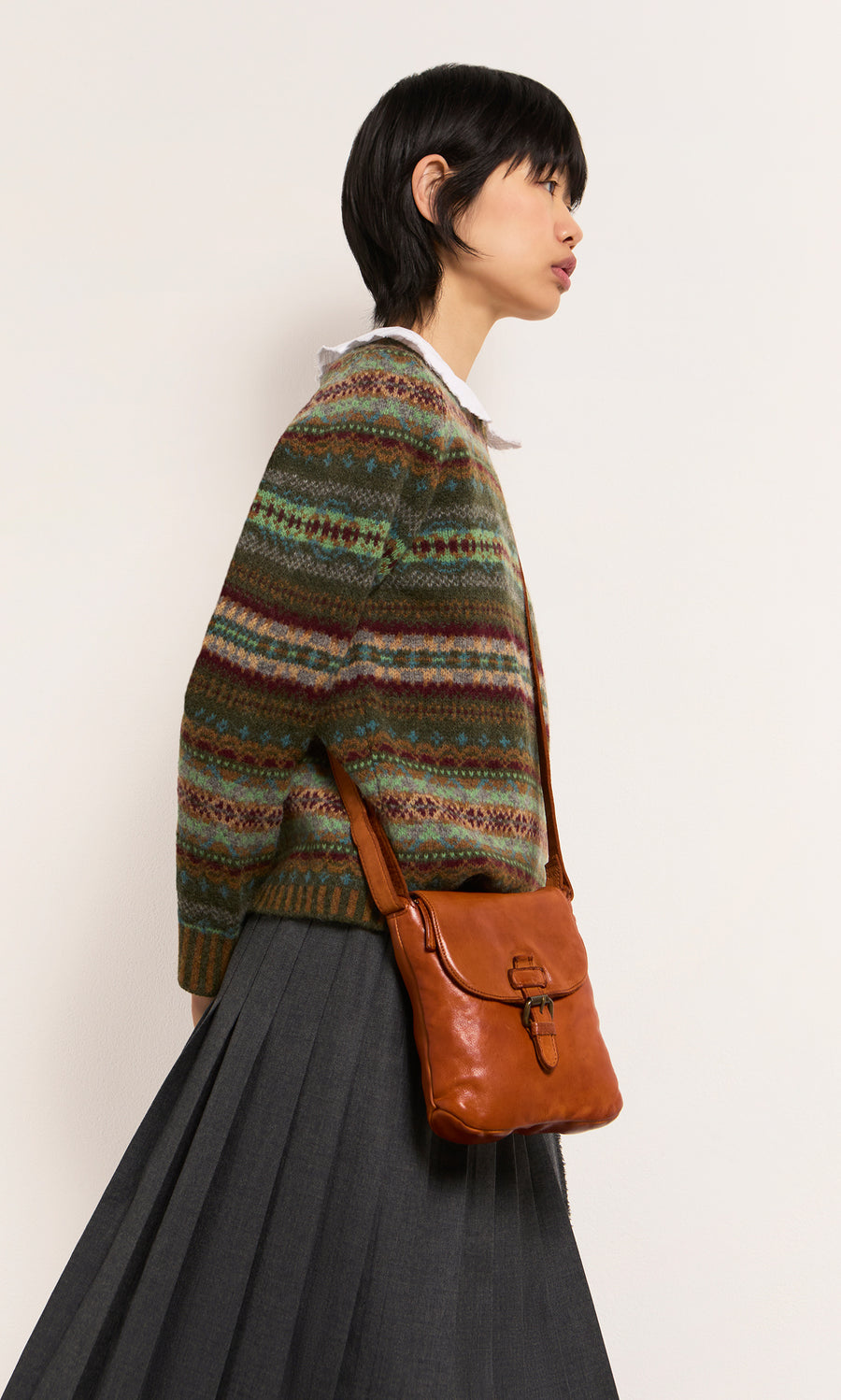 Plümo  Woman in a patterned sweater and gray skirt with a Parcel Bag - Chestnut stands against a plain backdrop.