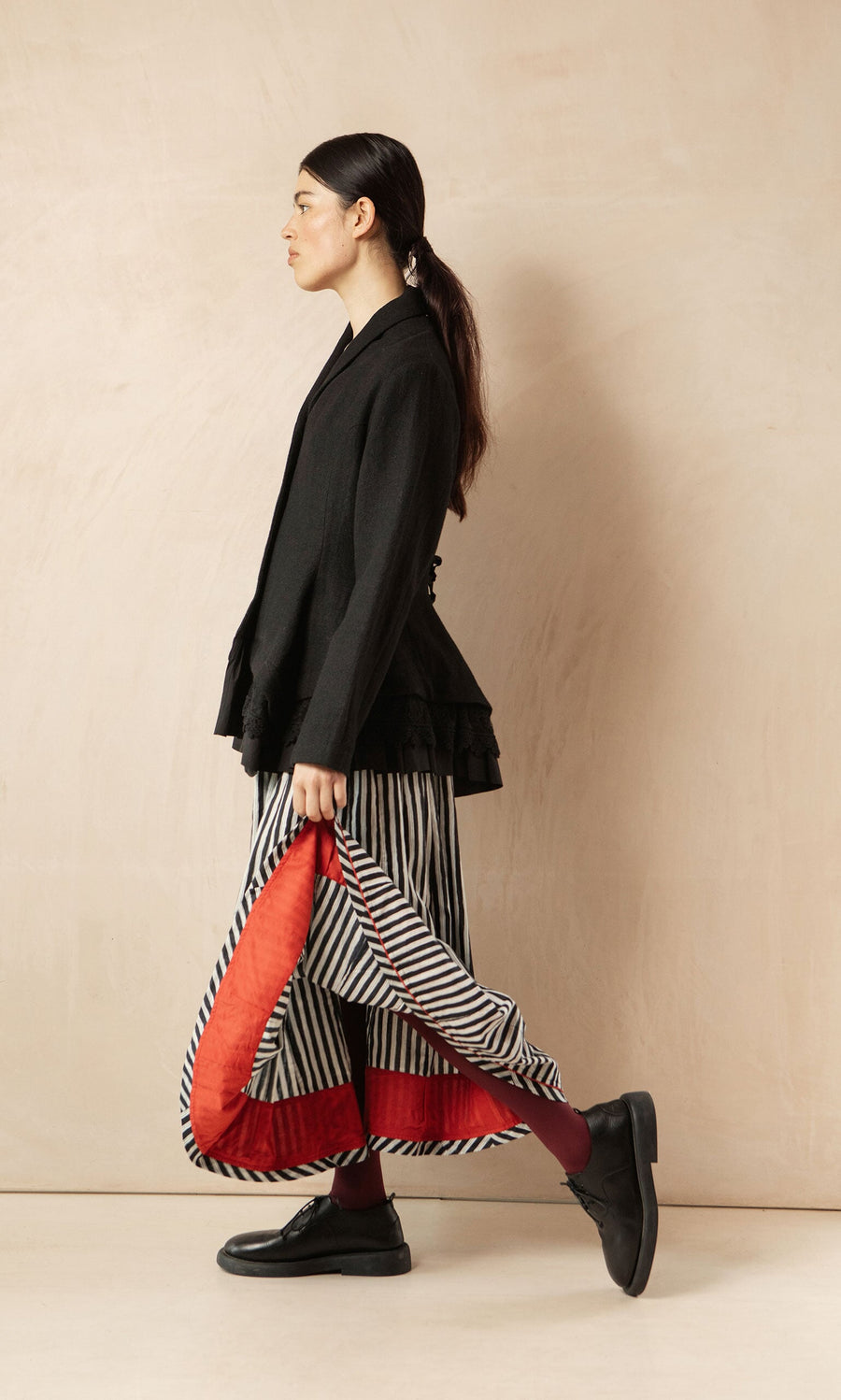 Plümo- Woman in profile wearing the Cara Riding Jacket, striped skirt, red tights, and black shoes against a neutral background.