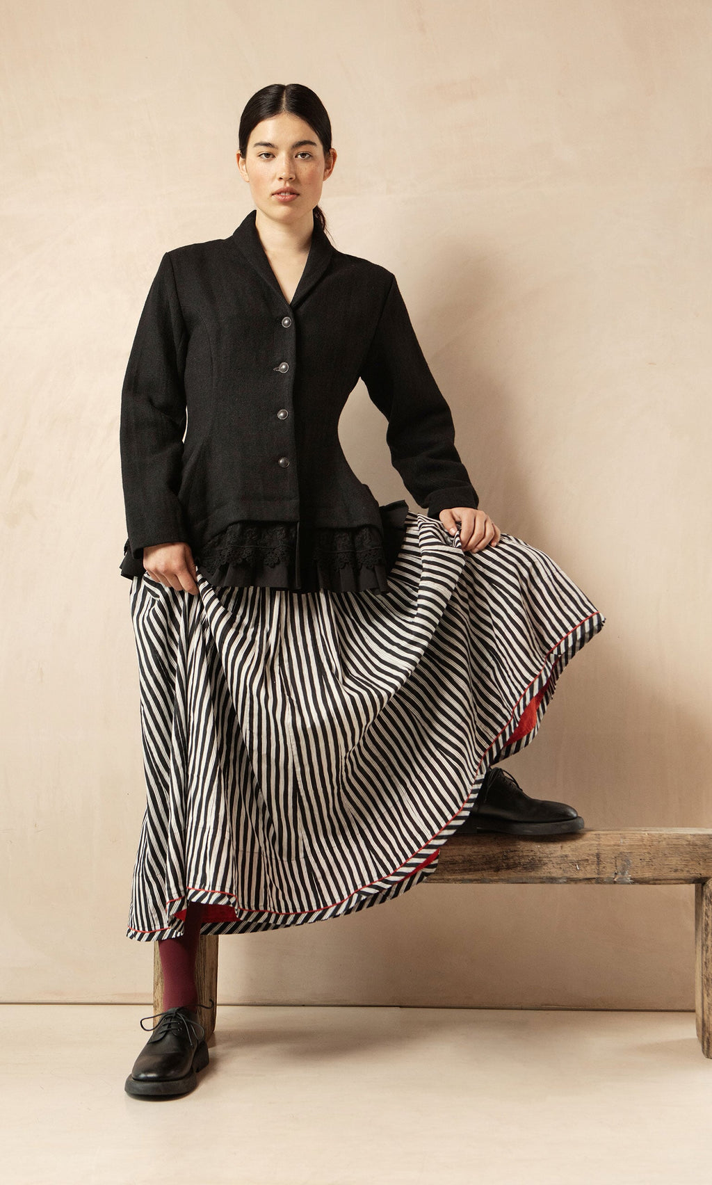 Plümo- A woman in the Cara Riding Jacket and a striped skirt stands on a bench, lifting her skirt to reveal red lining and black shoes.