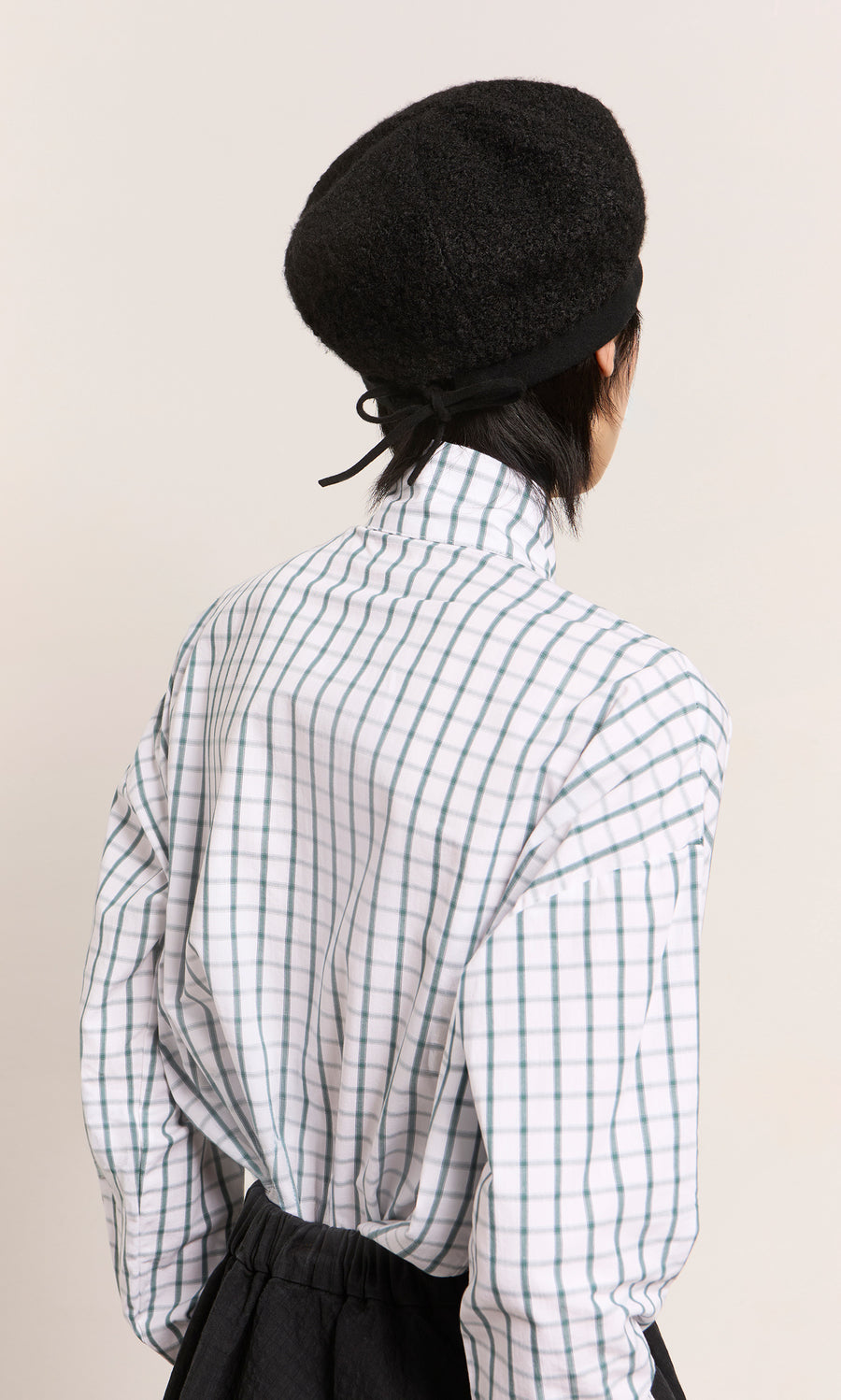 Plümo  Person facing away, wearing a black Perinne Beret and a white plaid high-collar shirt against a plain background.