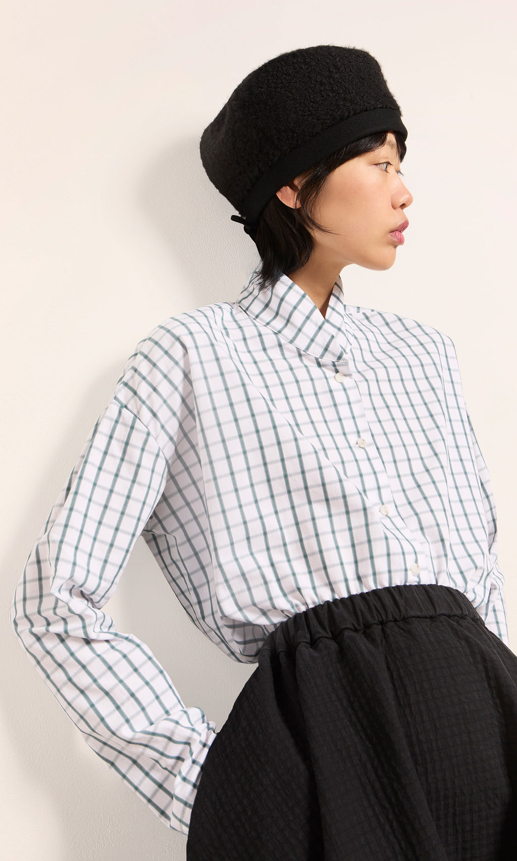 Plümo  Woman in a checkered shirt and black skirt wears the handmade Perinne Beret, gazing sideways against a plain backdrop.